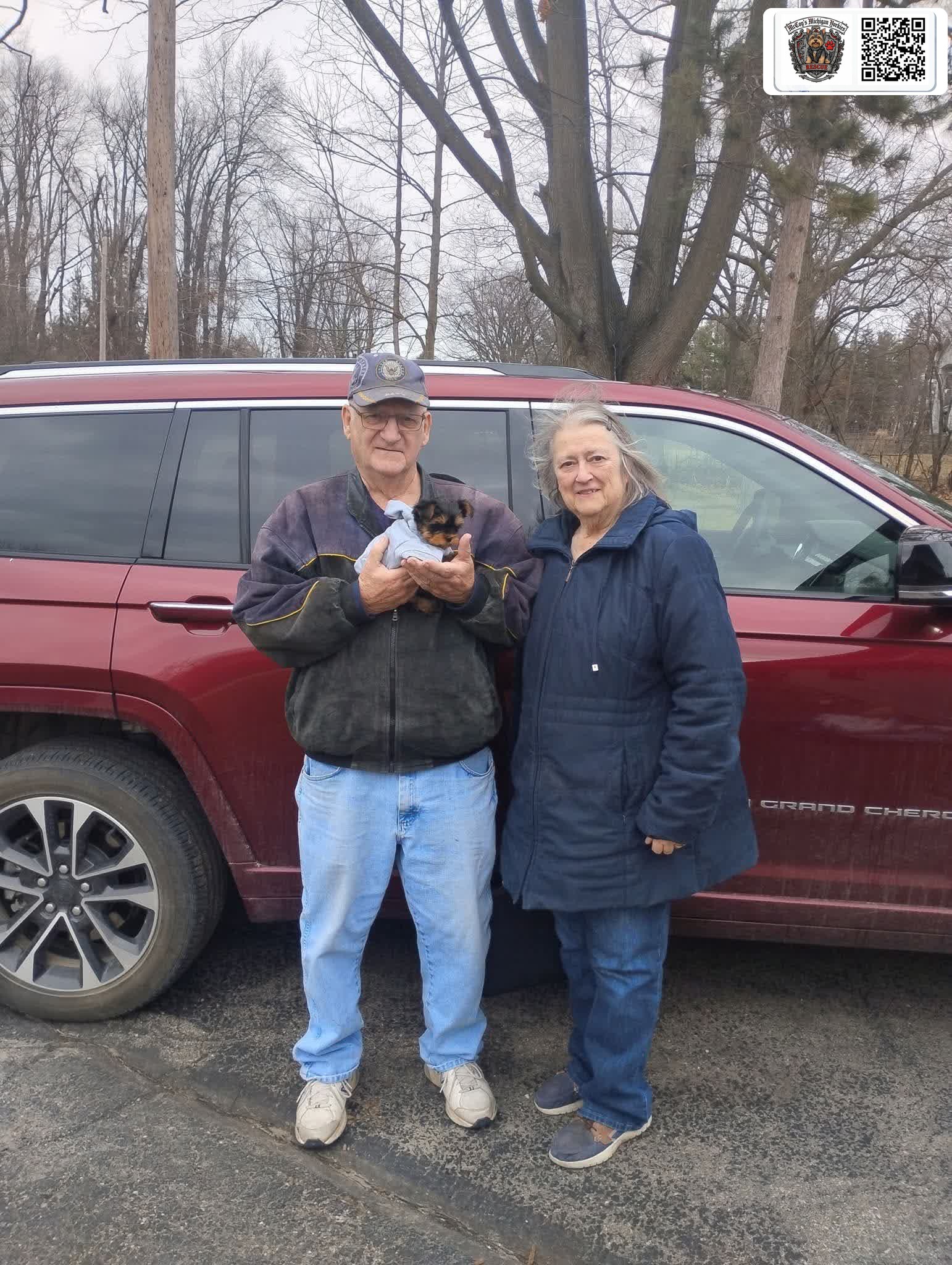 A returning McCoy's family smiling beside their red Jeep while holding their tiny new Yorkshire Terrier puppy